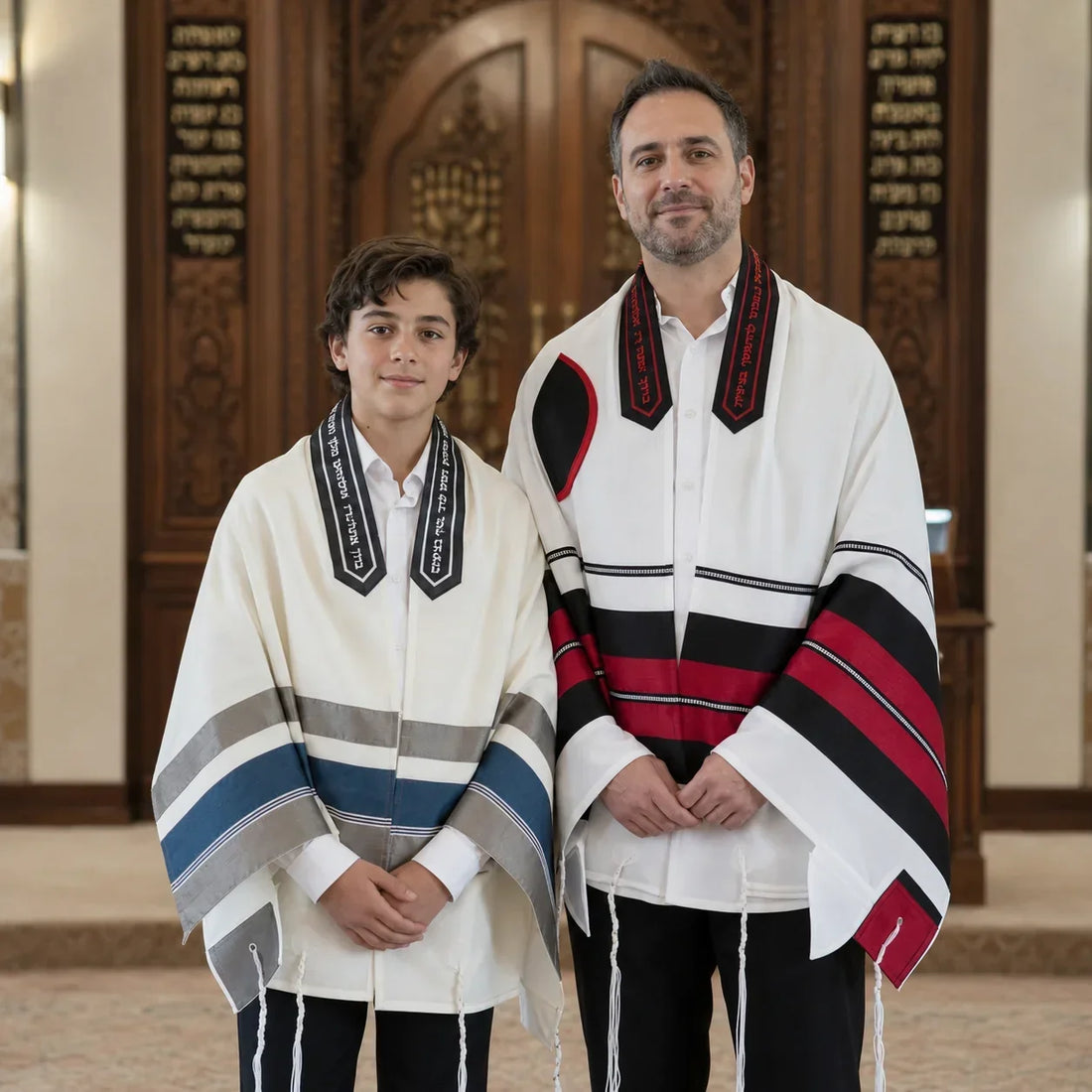 A father and son wearing traditional white Mitzvah Tallit with black, red, gray, and blue accents, standing together inside a synagogue.
